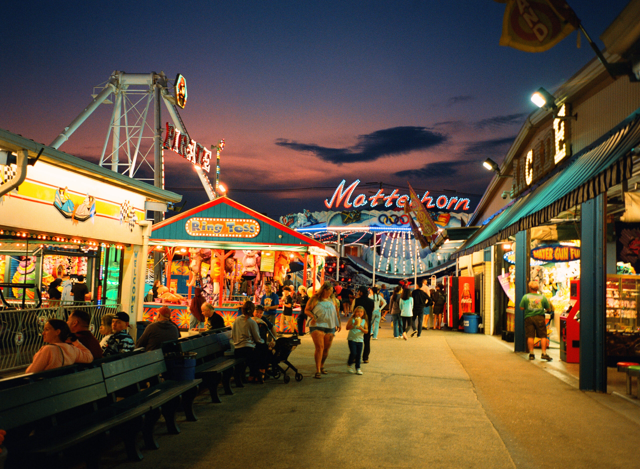 Old Orchard Beach Palace Playland wide neon scene with people milling about, arcade and Ring Toss booths, and the Matterhorn ride glowing in the background.